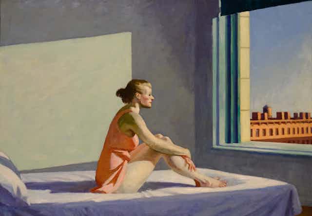A woman sits on a bed and stares out a window.