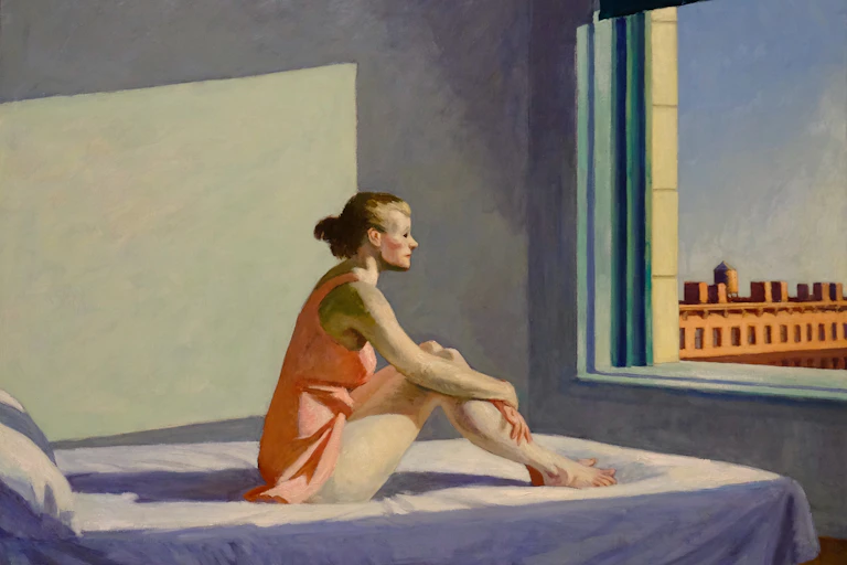 A woman sits on a bed and stares out a window.
