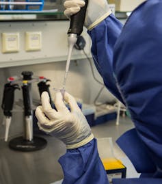 Someone wearing rubber gloves and a lab coat, placing a sample of DNA into a small test tube.