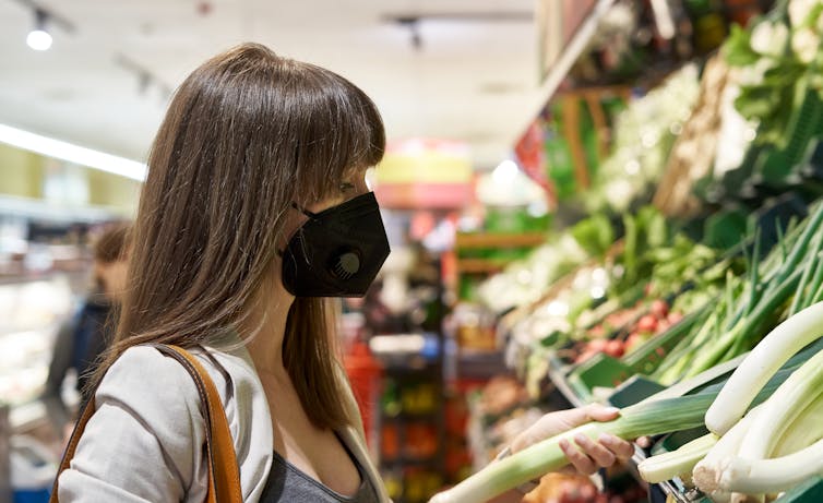 Woman with brown hair and black face mask picks up vegetable at supermarket