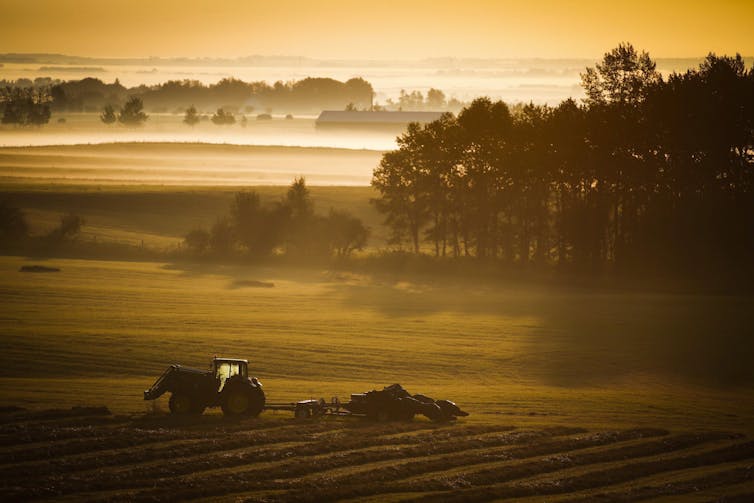 A farm tractor and baler sit in a hay field amid a misty sunrise.