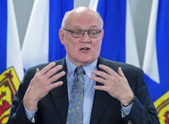 A man with glasses and wearing a suit gestures as he talks.