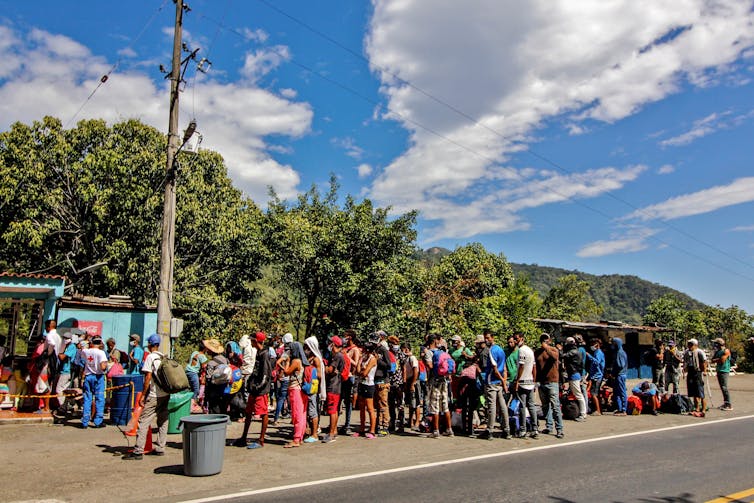 Migrants in a line wearing face masks in a verdant, warm-weather climate