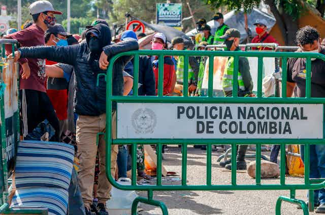 Line of people wearing face masks stand behind a gate that reads 'National Police of Colombia'