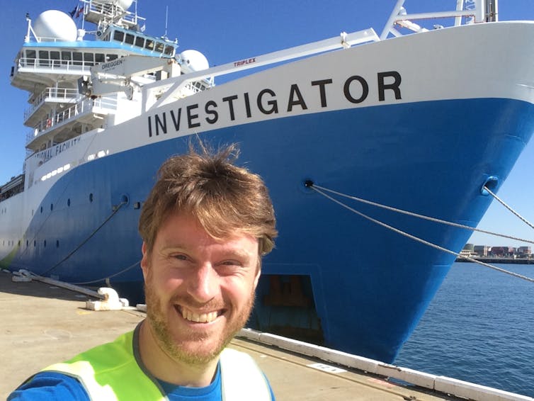 The author smiles in front of a blue and white ship, with 'Investigator' written on the side.