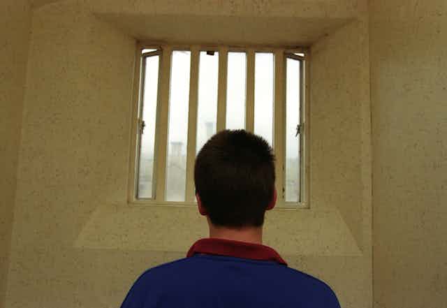 Man wearing blue top with back to camera looking out of window with bars on it