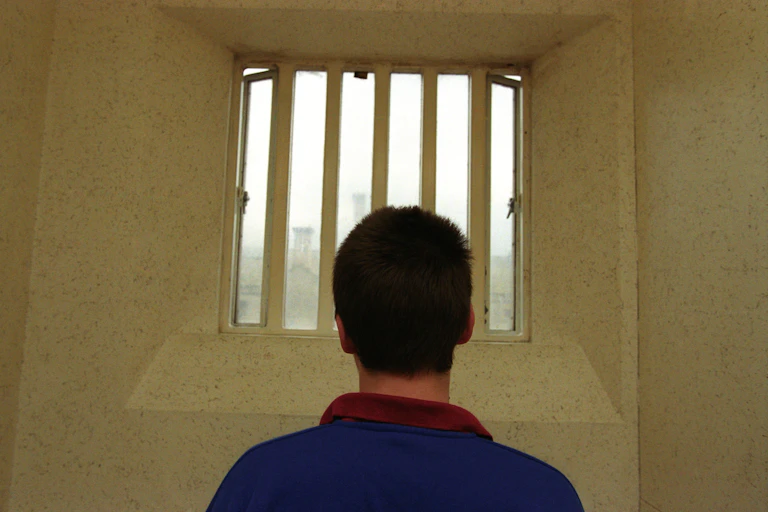Man wearing blue top with back to camera looking out of window with bars on it