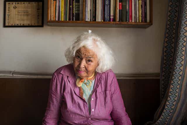 An elderly woman with white haircuts looking directly into camera, her head slightly cocked, a challenging look in her eyes. Behind her is a packed bookshelf on a wall.