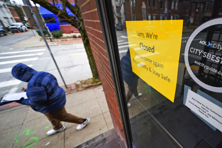 Person in winter clothing walking down a street in Pittsburgh, Pennsylvania, past a cafe with a sign reading, "Sorry, we're closed. Please come again. Keep healthy and stay safe."