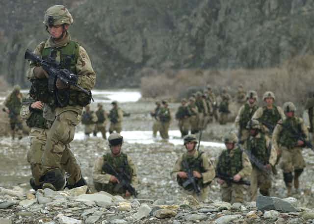 US troops carrying guns walking through a stream and on rocky ground in Afghanistan.