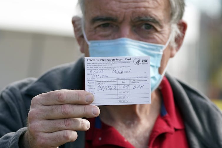 masked older man holds up his vaccination card