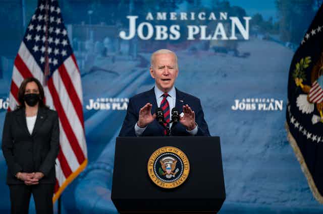 President Biden giving a speech in front of a sign that says "American Jobs Plan."