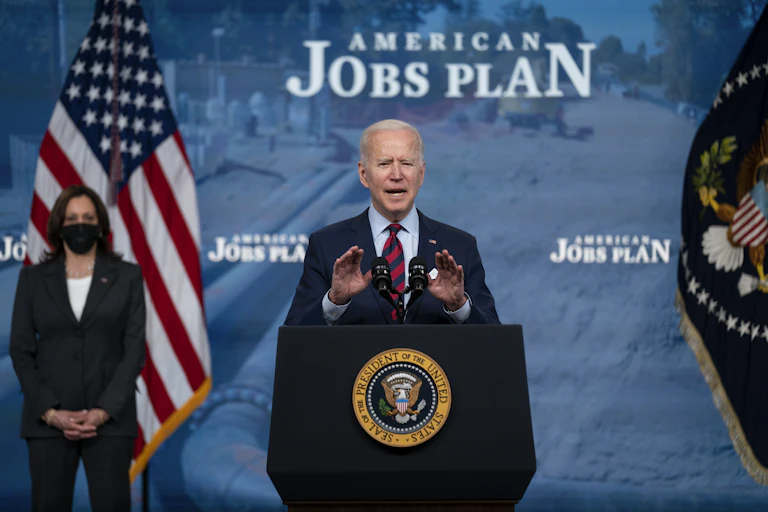 President Biden giving a speech in front of a sign that says "American Jobs Plan."