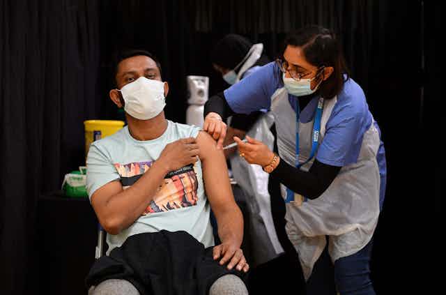 A woman administers a COVID-19 vaccination to a man seated with his sleeve rolled up