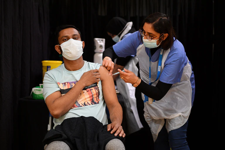 A woman administers a COVID-19 vaccination to a man seated with his sleeve rolled up