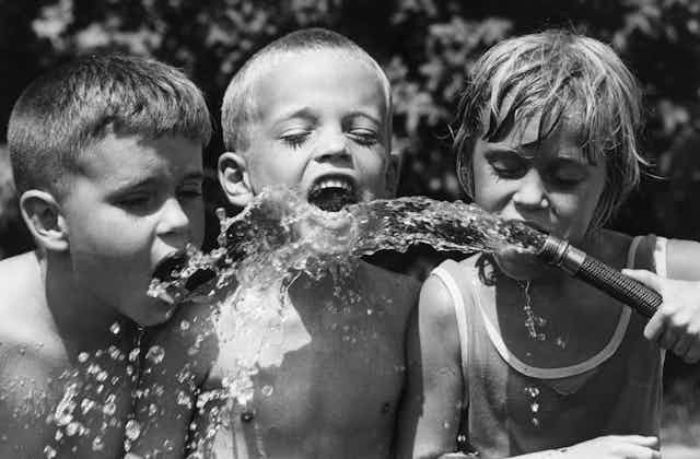 Three boys drink water from a hose in a black and white photo