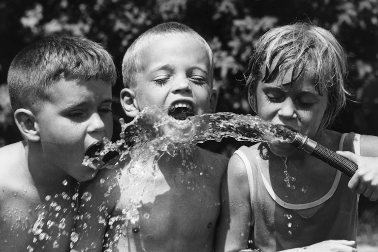 Three boys drink water from a hose in a black and white photo