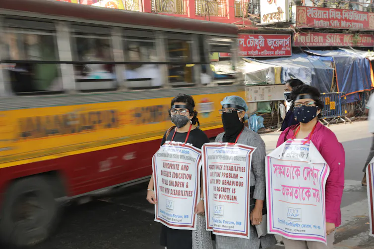Three people wearing face masks and holding protest banners stand in street as bus drives past