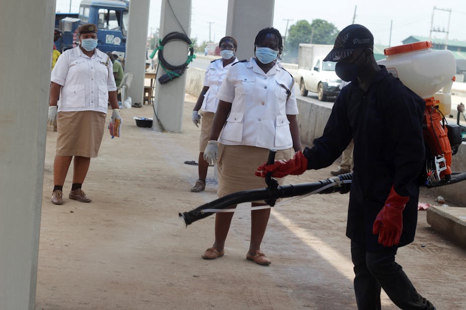 A man operates a chemical spray dispenser on a street, watched by women in uniform, all wearing protective masks and gloves