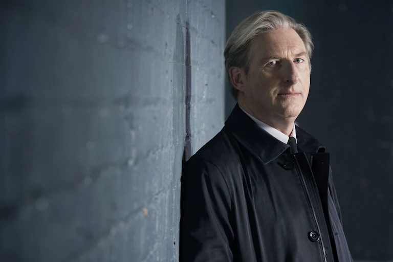 Photo of a man leaning up against a grey brick wall.