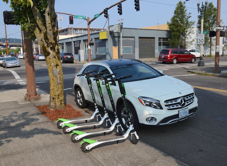 Cuatro scooters alineados al borde de una acera, cerca de la calle y un automóvil