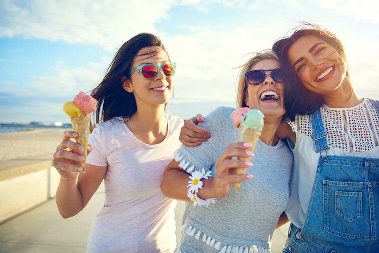 Des adolescentes rieuses mangent des cônes de glace en marchant le long d'une plage.