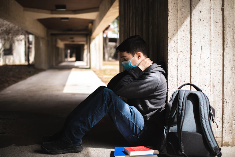 A young man wearing a mask sits underneath a bridge next to books and a backpack.
