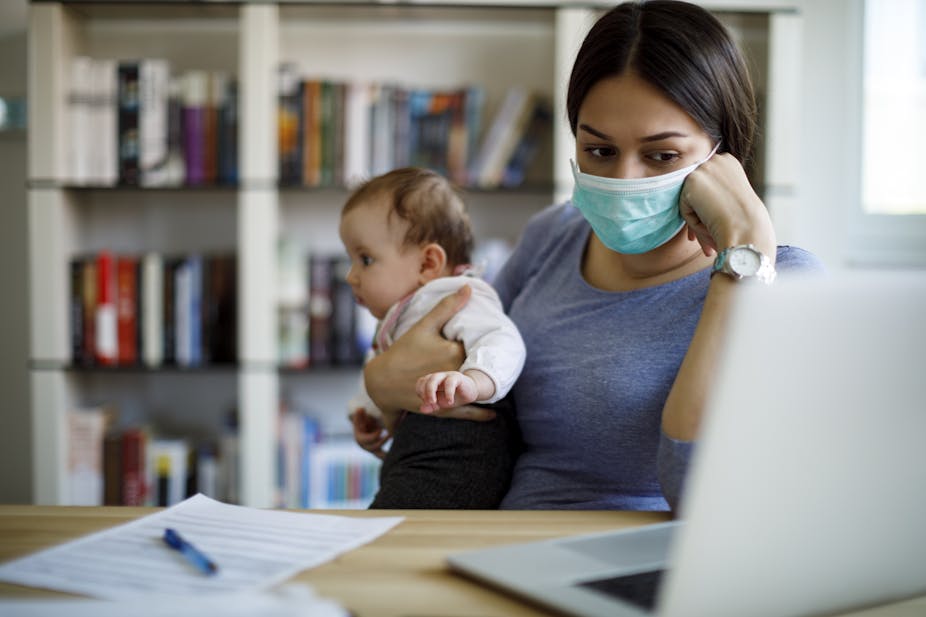 A woman in a surgical mask holds a baby while looking at a laptop and paperwork