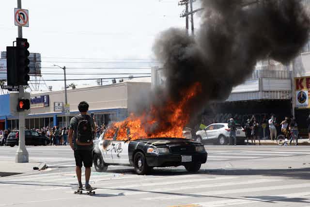 A skateboarder films a burning cop car in an intersection