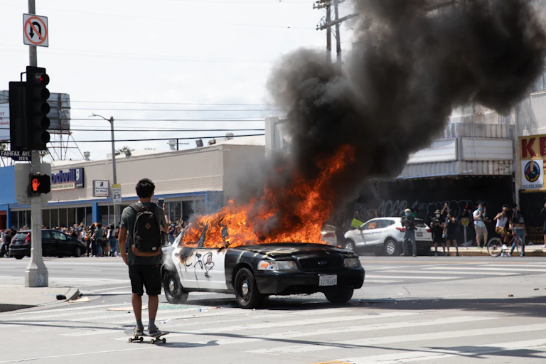 A skateboarder films a burning cop car in an intersection