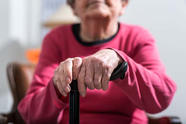 An elderly woman's hands rest on a walking stick.