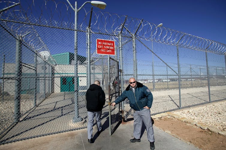 Guard opens the gate for a person in a high chain link fence topped with barbed wire