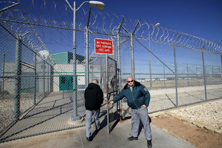 Guard opens the gate for a person in a high chain link fence topped with barbed wire