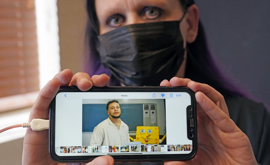 Carrie Shipp is wearing a black surgical mask and looking at the camera. She holds up a phone with a picture of her 21-year-old son, Matthew Shipp, who is imprisoned. 