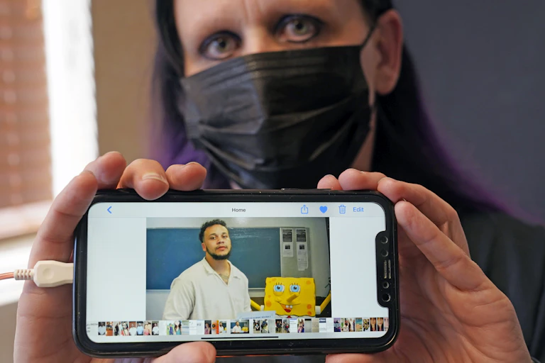 Carrie Shipp is wearing a black surgical mask and looking at the camera. She holds up a phone with a picture of her 21-year-old son, Matthew Shipp, who is imprisoned.