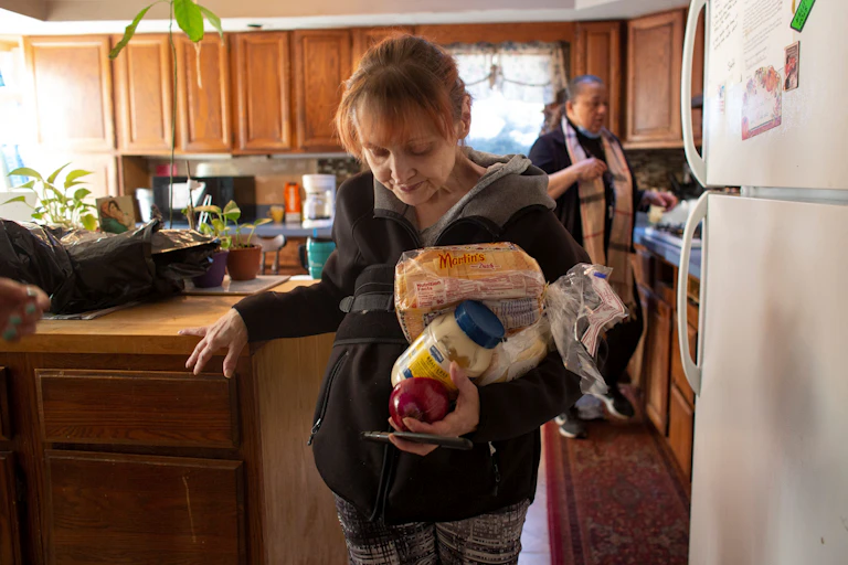 Two women in a kitchen, one holding bread, a mayonnaise jar and an apple