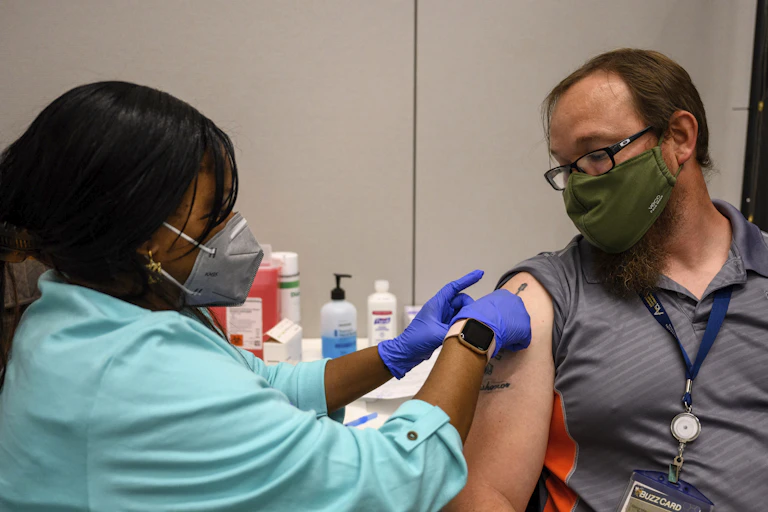 A female nurse wearing blue gloves gives a bespectacled man a Pfizer COVID-19 shot on the campus of Georgia Tech