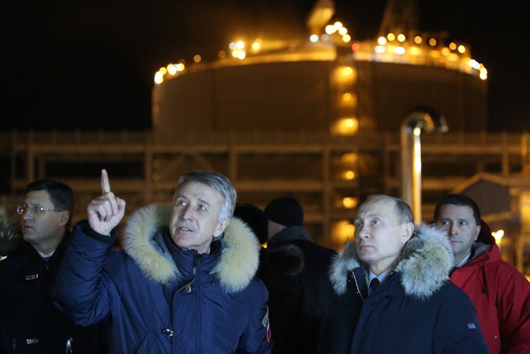 Russian President Vladimir Putin stands a man who is pointing outside the lit-up LNG plant.