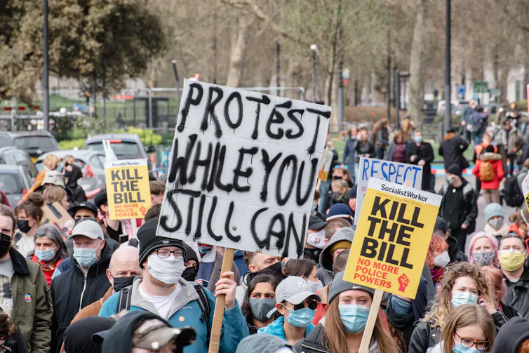 Crowd of protesters wearing face masks hold signs that say 'Protest while you still can' and 'kill the bill'