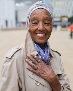 An aging woman in a trench coat, colourful scarf and head scarf.