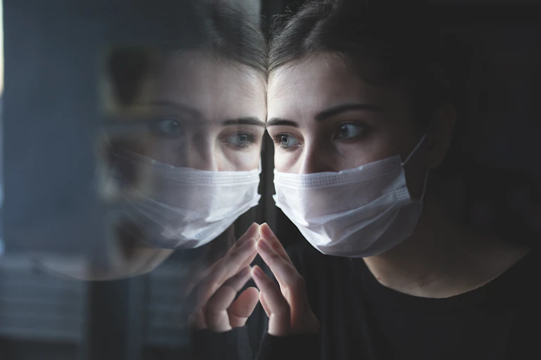 A young woman wearing a mask stares out a window.