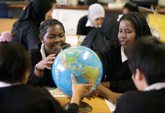 Two black schoolgirls with braids sit in front of two other schoolgirls wearing hijabs 