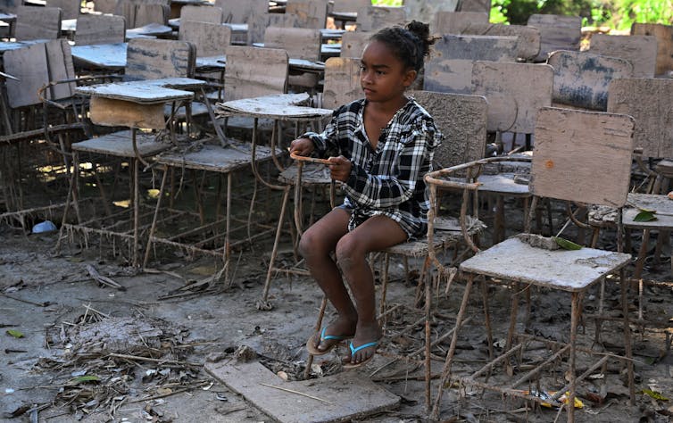 Una niña sentada en una silla escolar embarrada y destruida en un terreno embarrado y desordenado