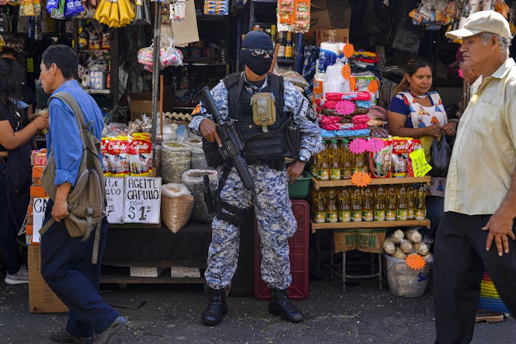 Un oficial de policía con equipo SWAT completo con ametralladoras se encuentra fuera de una pequeña tienda en una calle de la ciudad mientras la gente pasa caminando