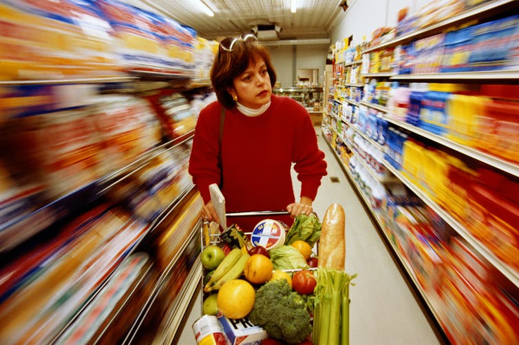 A stressed woman pushes a cart down a supermarket aisle.