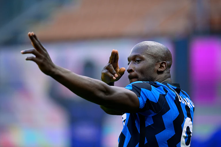 A man with a shaven head in a football jersey raises his arms, almost crossing them, fingers pointing in two different directions as if he's holding an invisible bow and arrow.