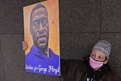 A woman in a pink mask holds up a poster of George Floyd.
