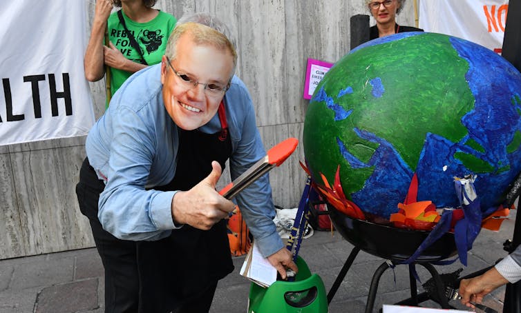 Person in Scott Morrison mask leans over burning globe, holding tongs