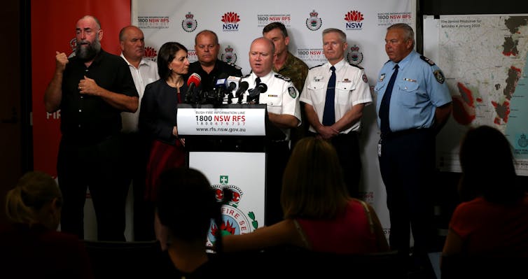An Auslan interpreter stands next to politicians and emergency officials during a TV broadcast.