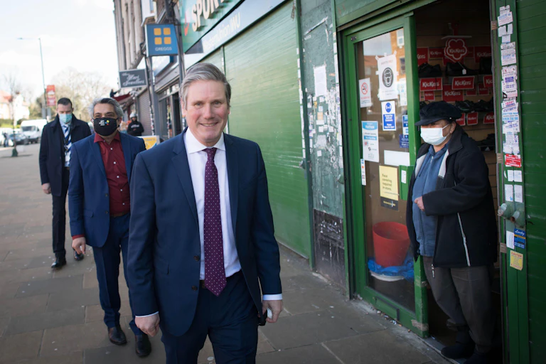 Keir Starmer walking past a shop as a person wearing a mask looks on.